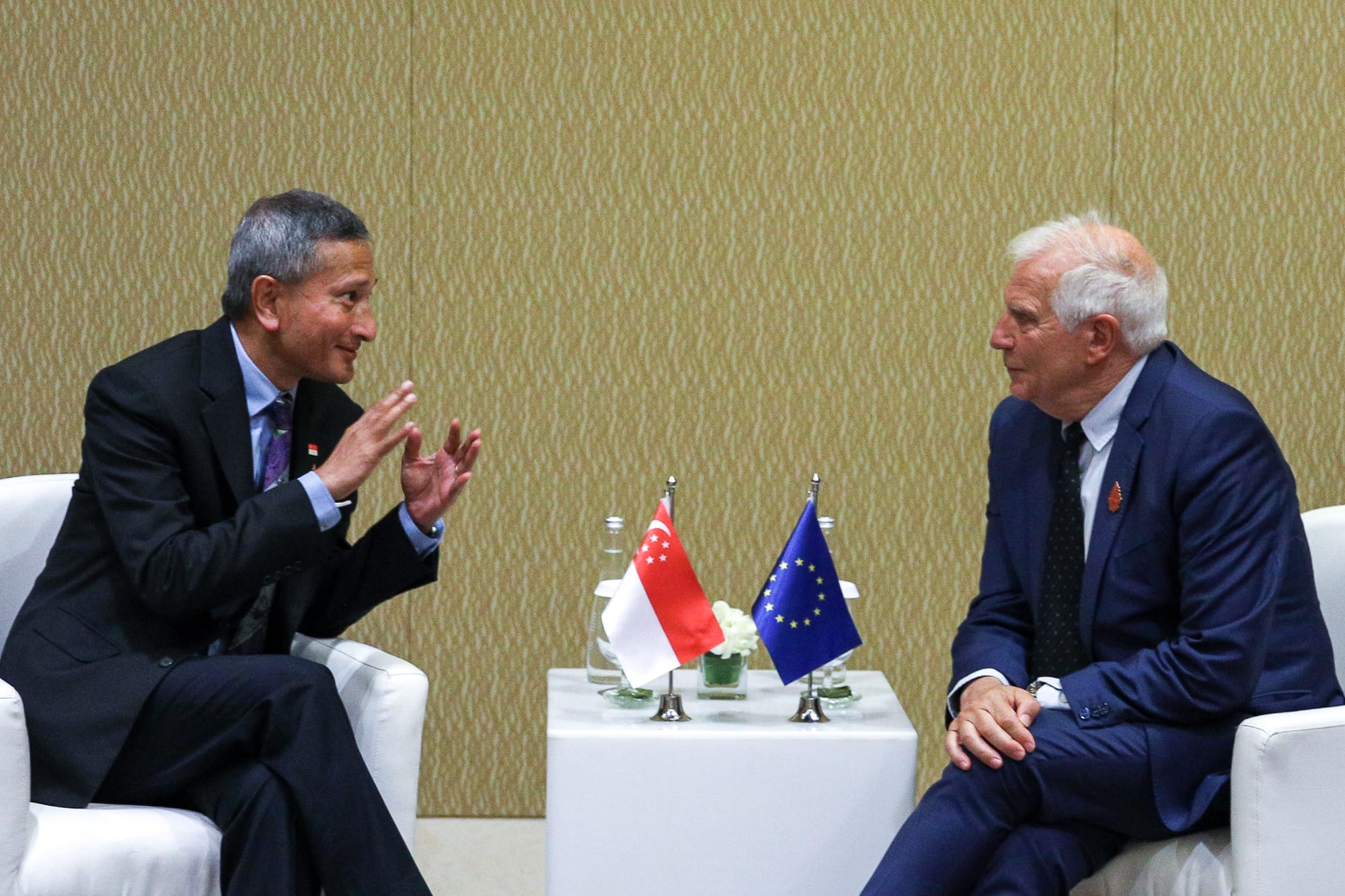 Two men in suits seated facing each other with aide behind; mics, documents, snacks on tables.